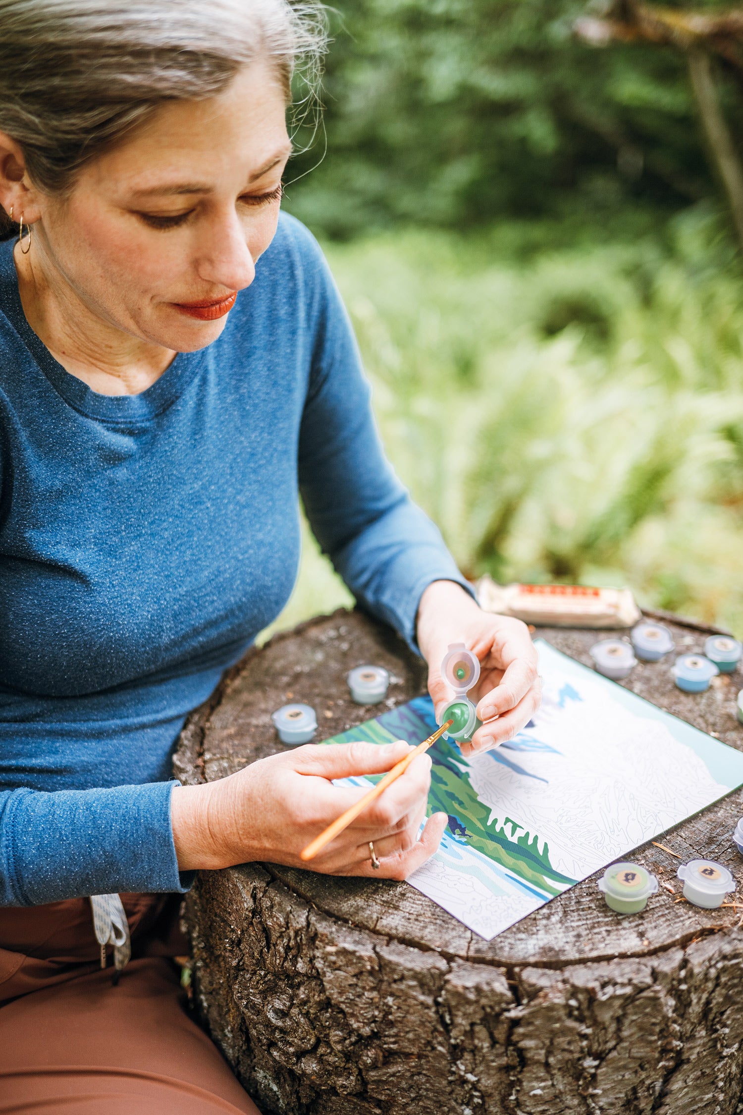 Woman in blue shirt painting a Denali National Park paint-by-number kit outdoors with a tree stump as a table.