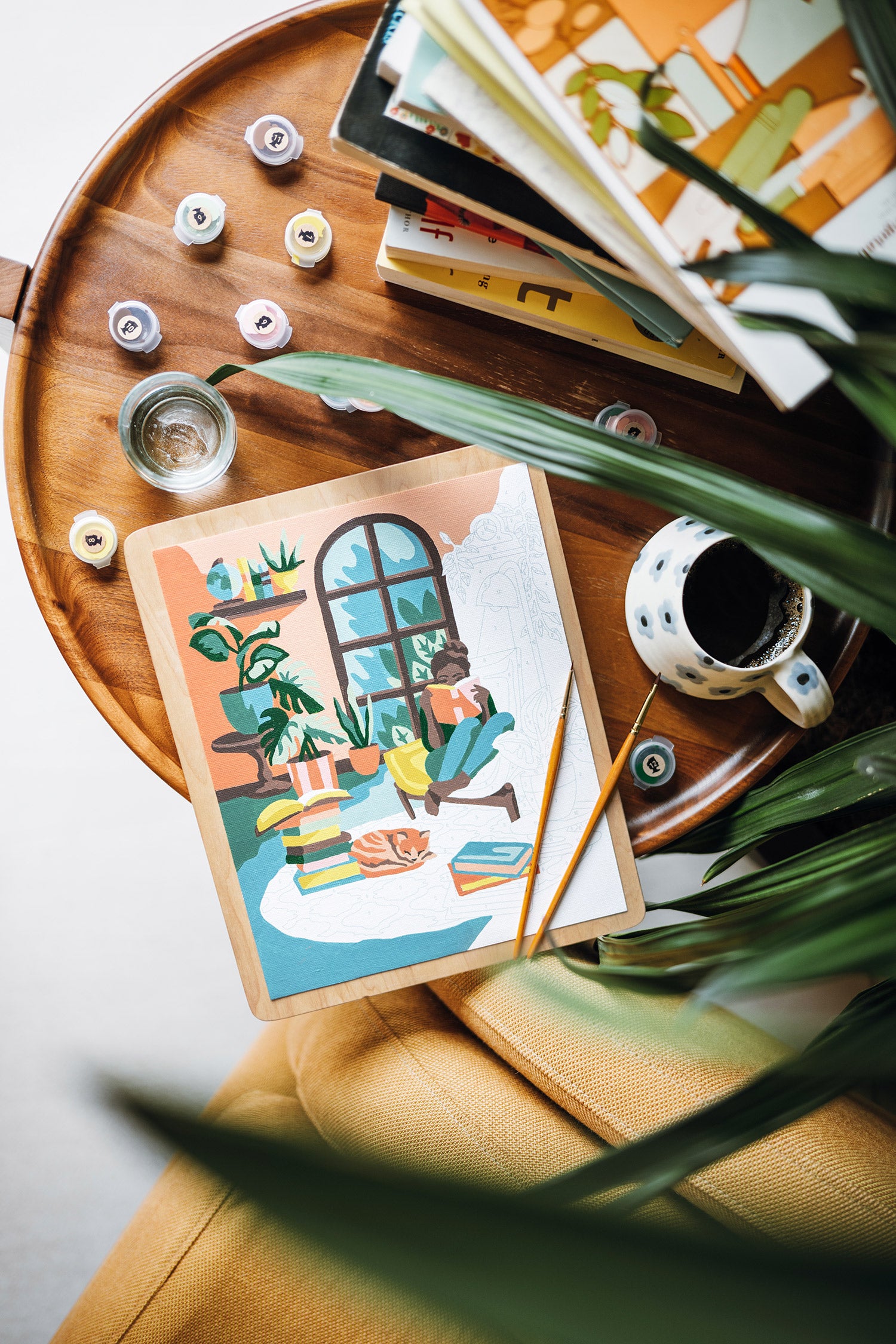 Wooden side table with A Novel Afternoon paint-by-number kit, coffee cup, and books beside a yellow chair and plants.