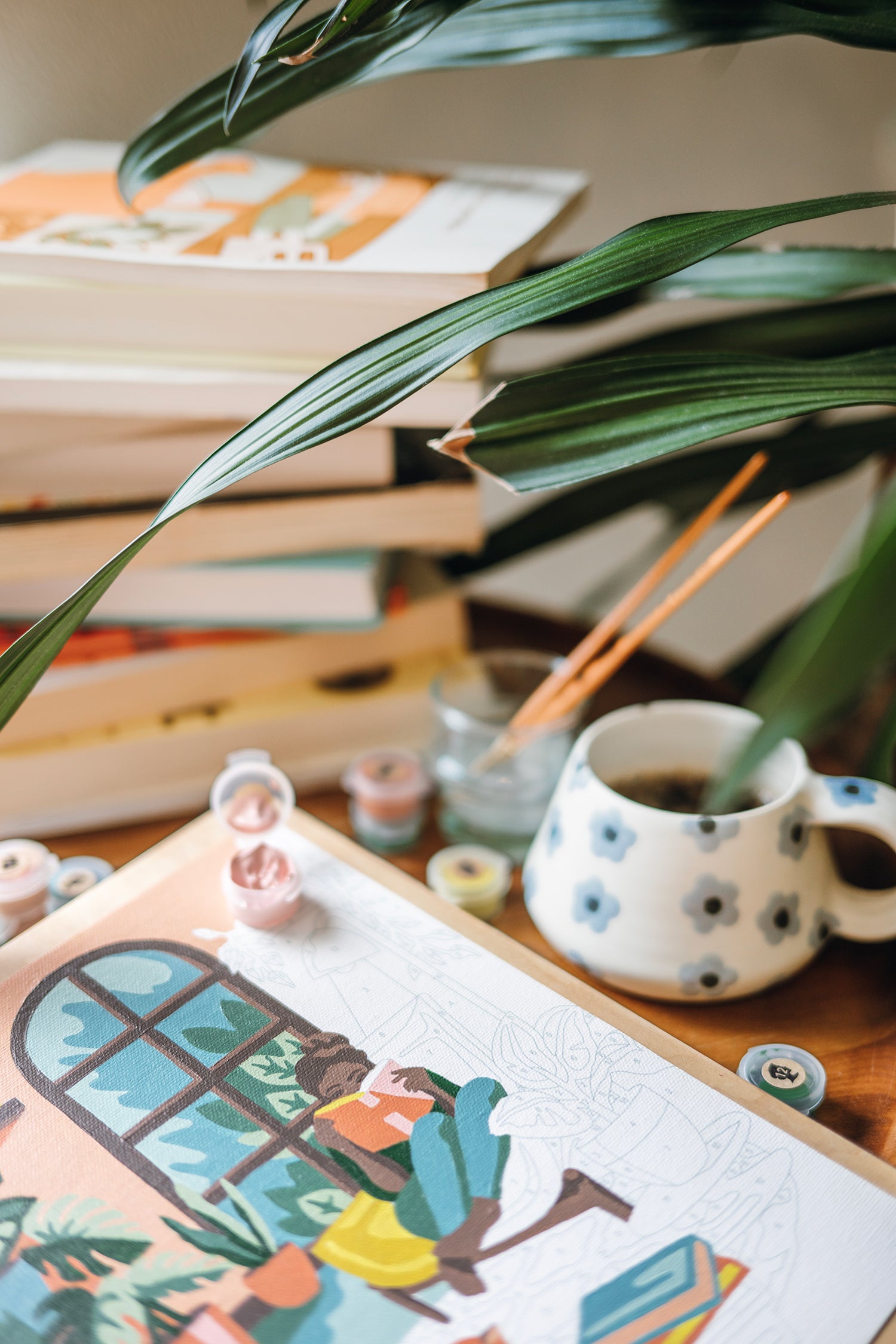Wooden side table with A Novel Afternoon paint-by-number kit, coffee cup, and books beside a yellow chair and plants.