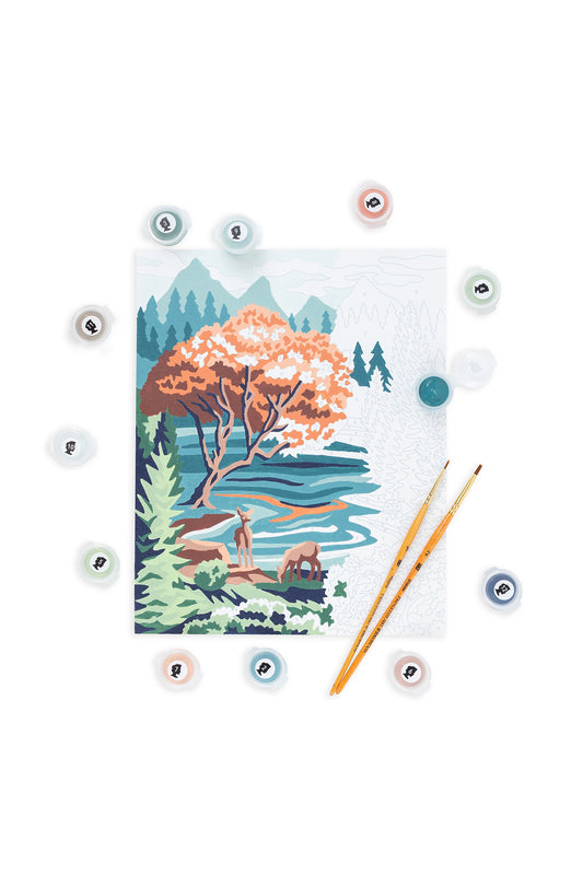 Partially painted Pine & Lake paint-by-number canvas on a white background, surrounded by numbered paint pots and two paintbrushes. The artwork shows a Pacific Northwest lake scene with pine trees, an autumn-colored tree, flowing water, and deer, with some sections still unpainted.