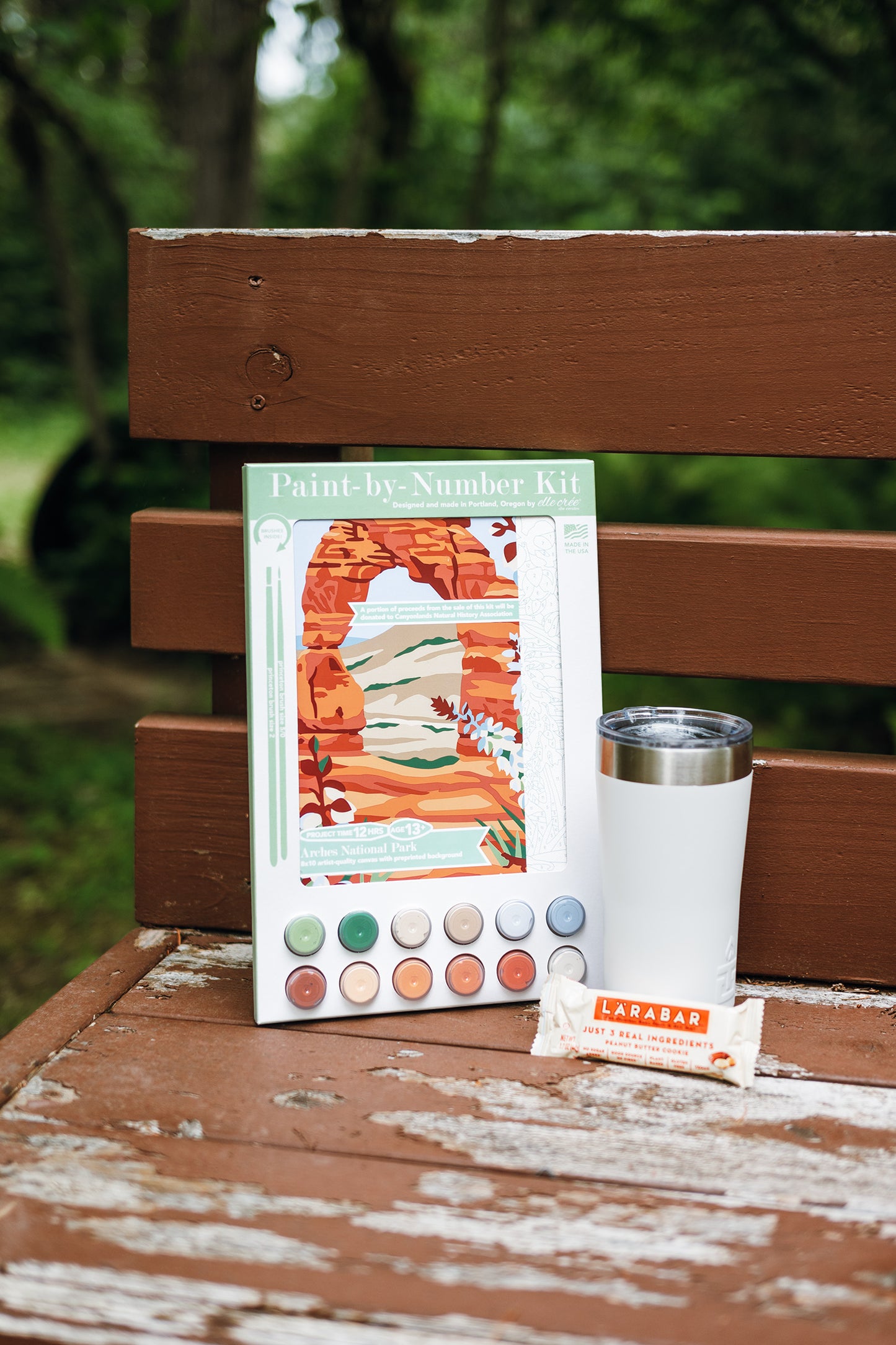 Arches National Park Paint-by-Number kit with a protein bar and cup on a wooden bench outdoors.