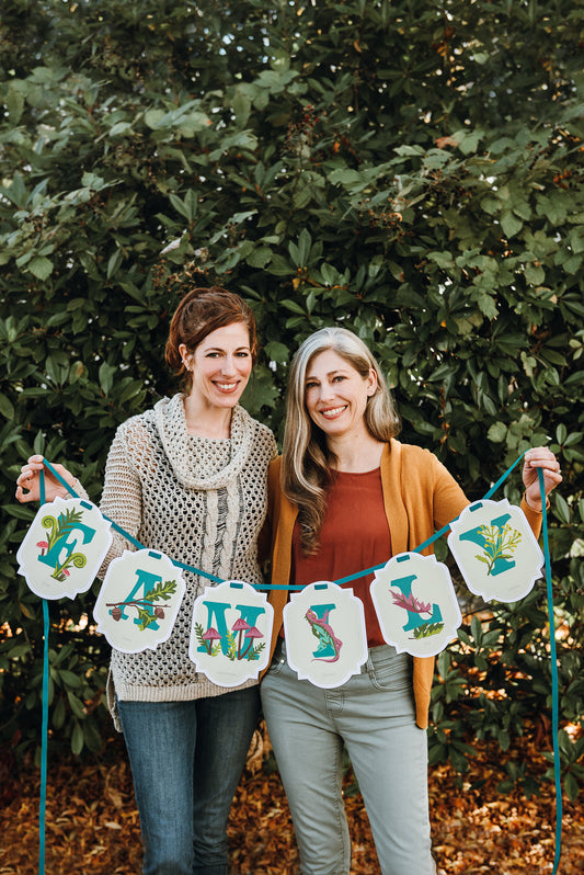 Two women holding up a fully painted paint-by-number banner kit that reads "Family."