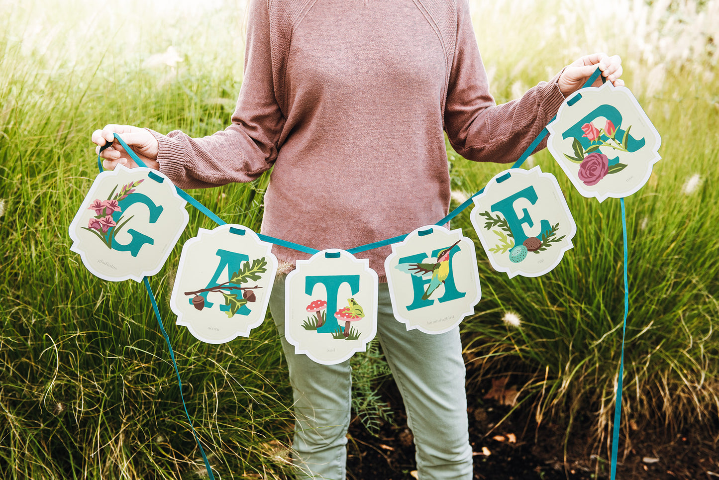 Person holding a paint-by-number banner with floral and bird designs that reads GATHER in a grassy outdoor setting.