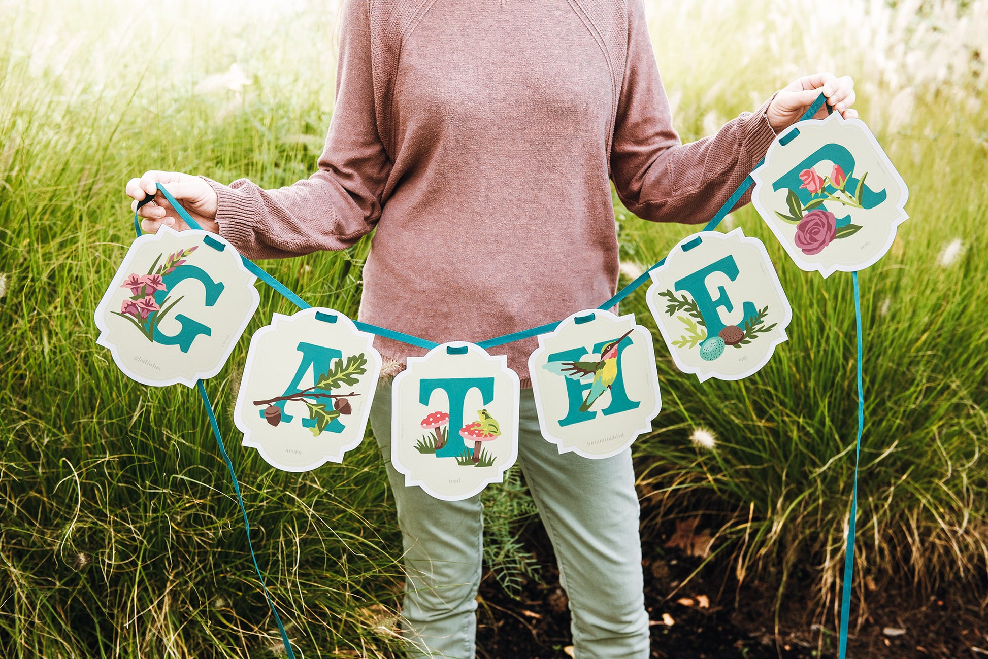 Person holding a paint-by-number banner with floral and bird designs that reads GATHER in a grassy outdoor setting.