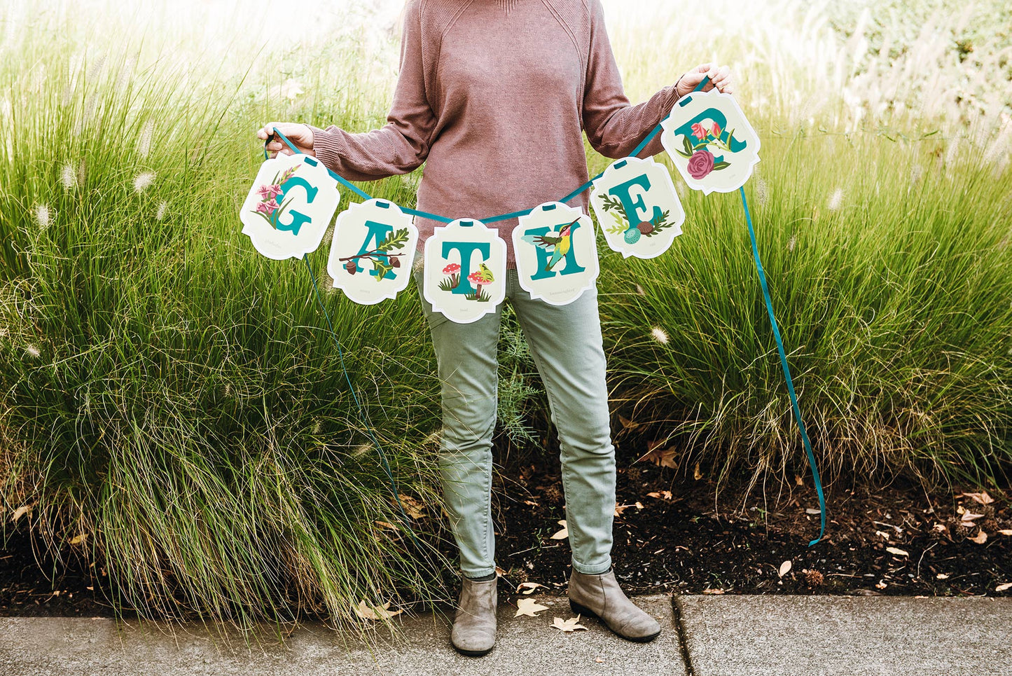 Person holding a paint-by-number banner with floral and bird designs that reads GATHER in a grassy outdoor setting.