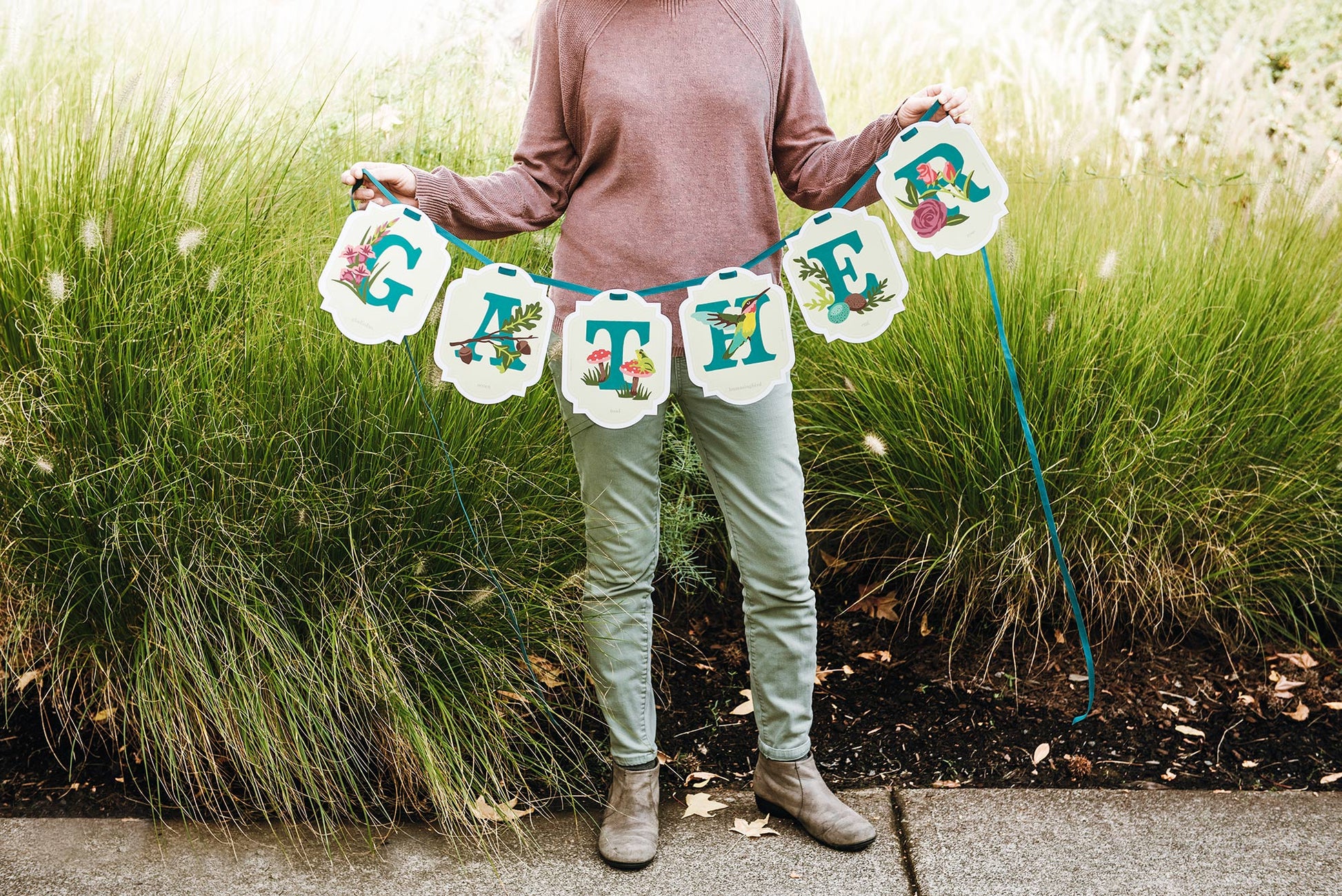 Person holding a paint-by-number banner with floral and bird designs that reads GATHER in a grassy outdoor setting.