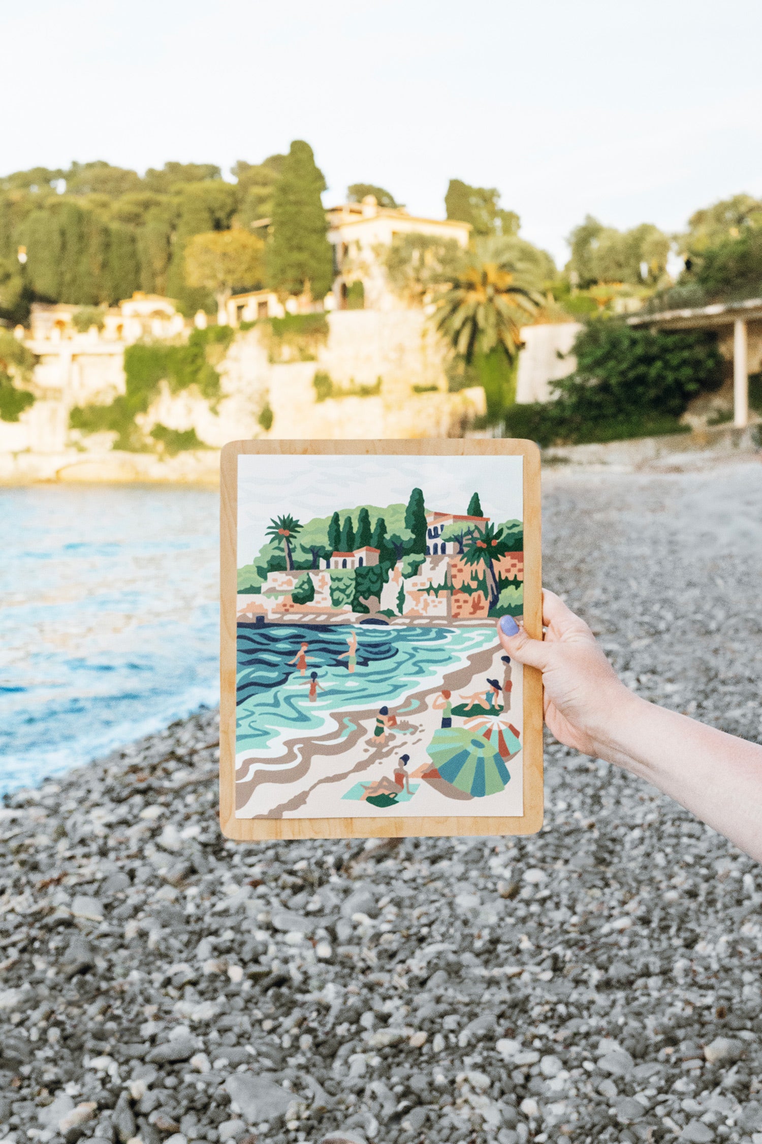 Finished Paint-by-Number kit of Paloma Beach held up in front of the actual Paloma Beach in the South of France.