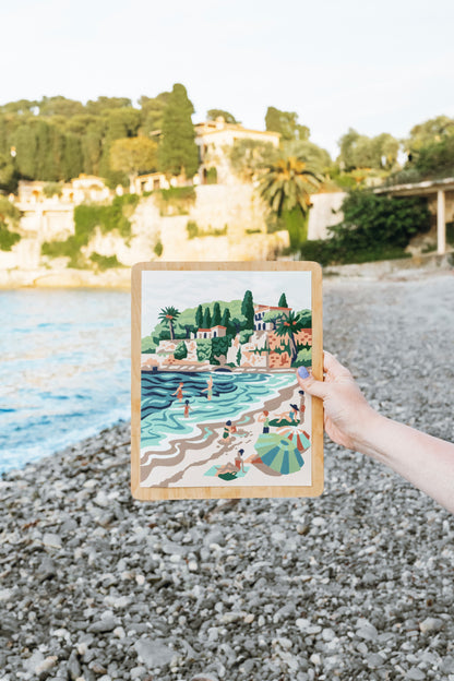 Finished Paint-by-Number kit of Paloma Beach held up in front of the actual Paloma Beach in the South of France.