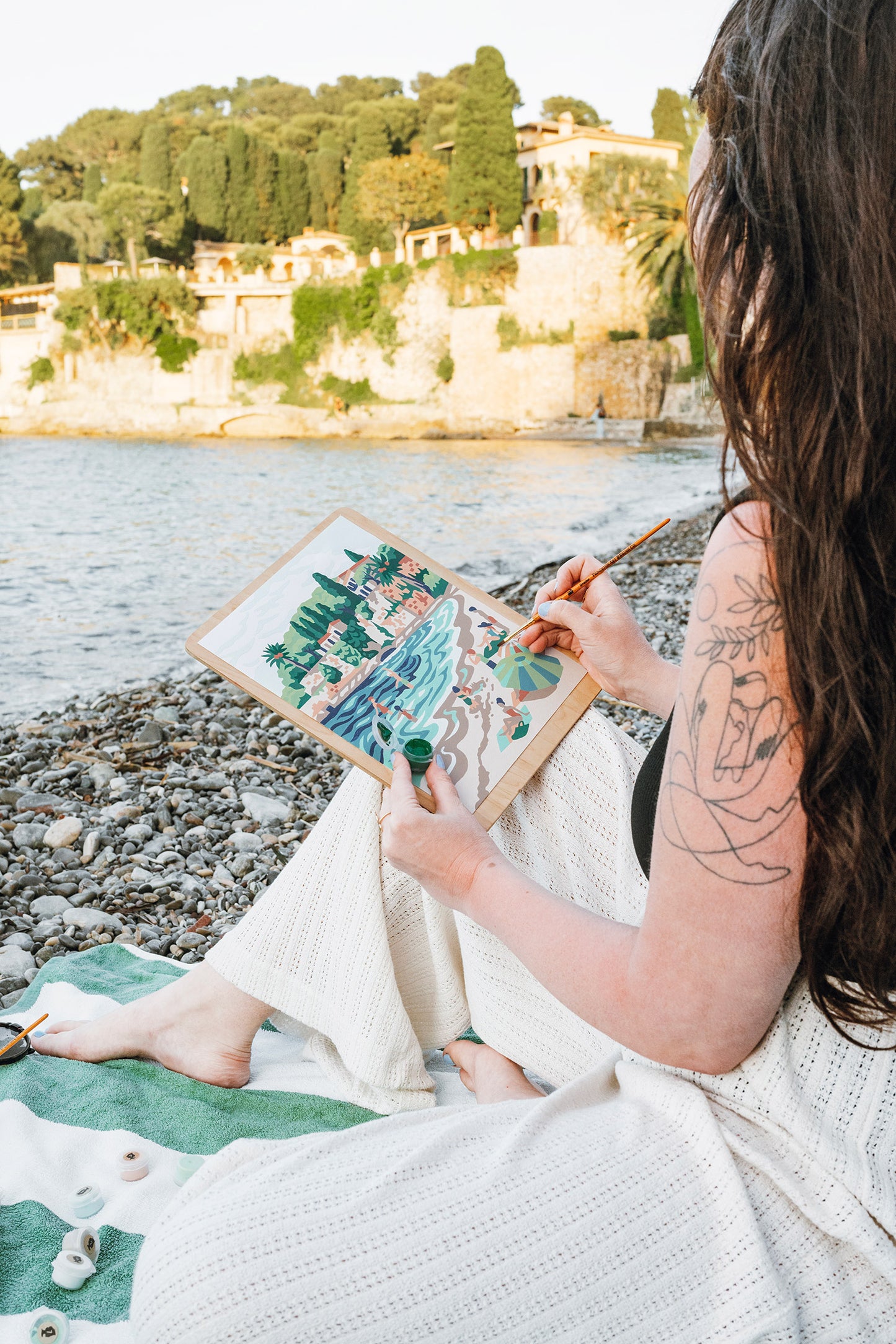 Woman painting a paint-by-number kit of Paloma Beach while sitting on the shore of Paloma Beach in the South of France.