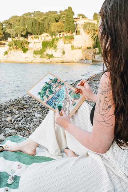 Woman painting a paint-by-number kit of Paloma Beach while sitting on the shore of Paloma Beach in the South of France.