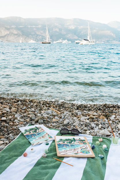 Riviera Romp (Paloma Beach) paint-by-number kit sitting scattered on a towel on the pebble beach in St. Jean Cap Ferrat France.