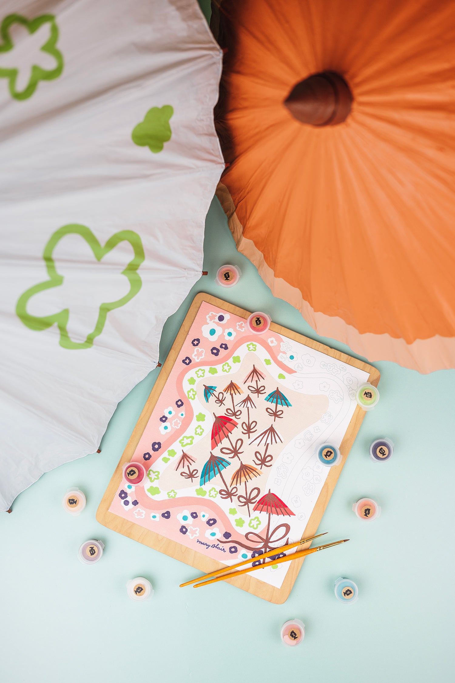 Mary Blair Parasols paint-by-number kit lying on a tabletop beside painted parasols.