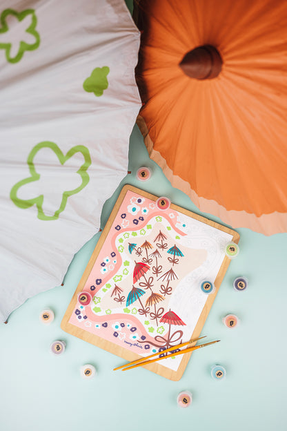 Mary Blair Parasols paint-by-number kit lying on a tabletop beside painted parasols.