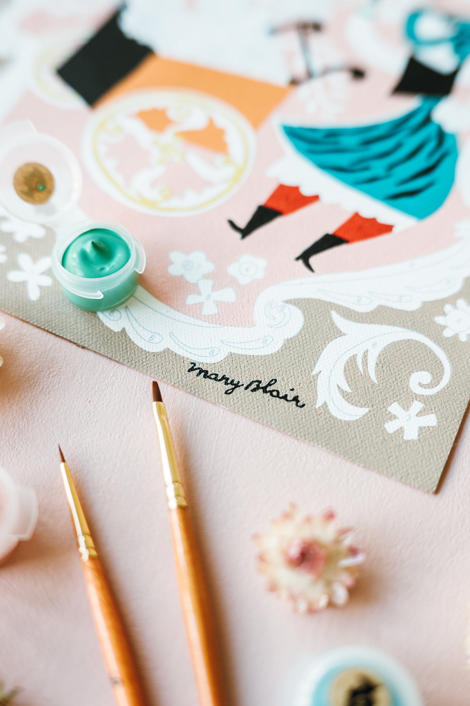 Close-up of Mary Blair’s Petal Peddler paint-by-number kit with artist’s signature, paint pot, and brushes on pink surface.