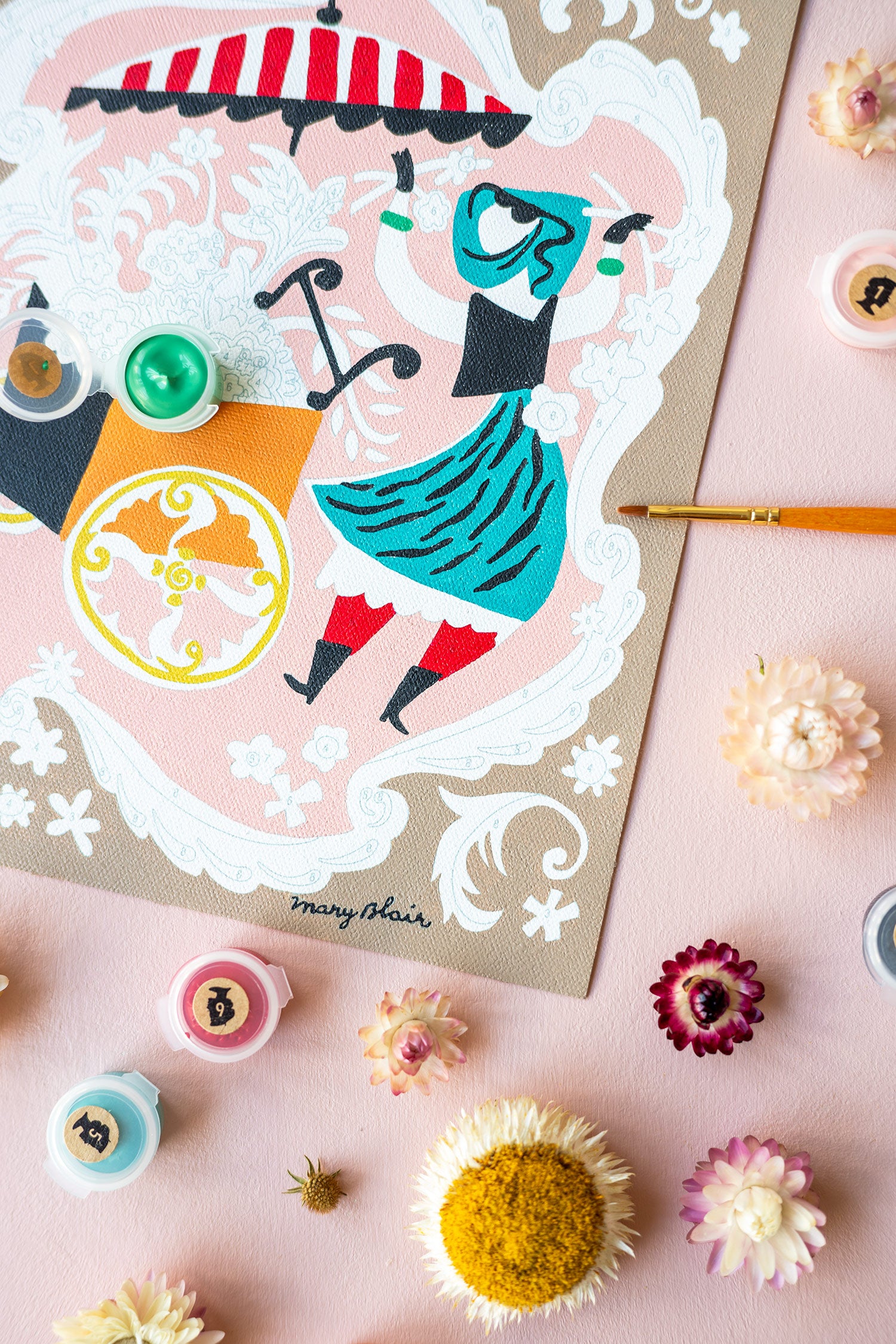 Mary Blair’s Petal Peddler paint-by-number kit in progress with flower cart scene, paint pots, brushes, and dried flowers on pink background.