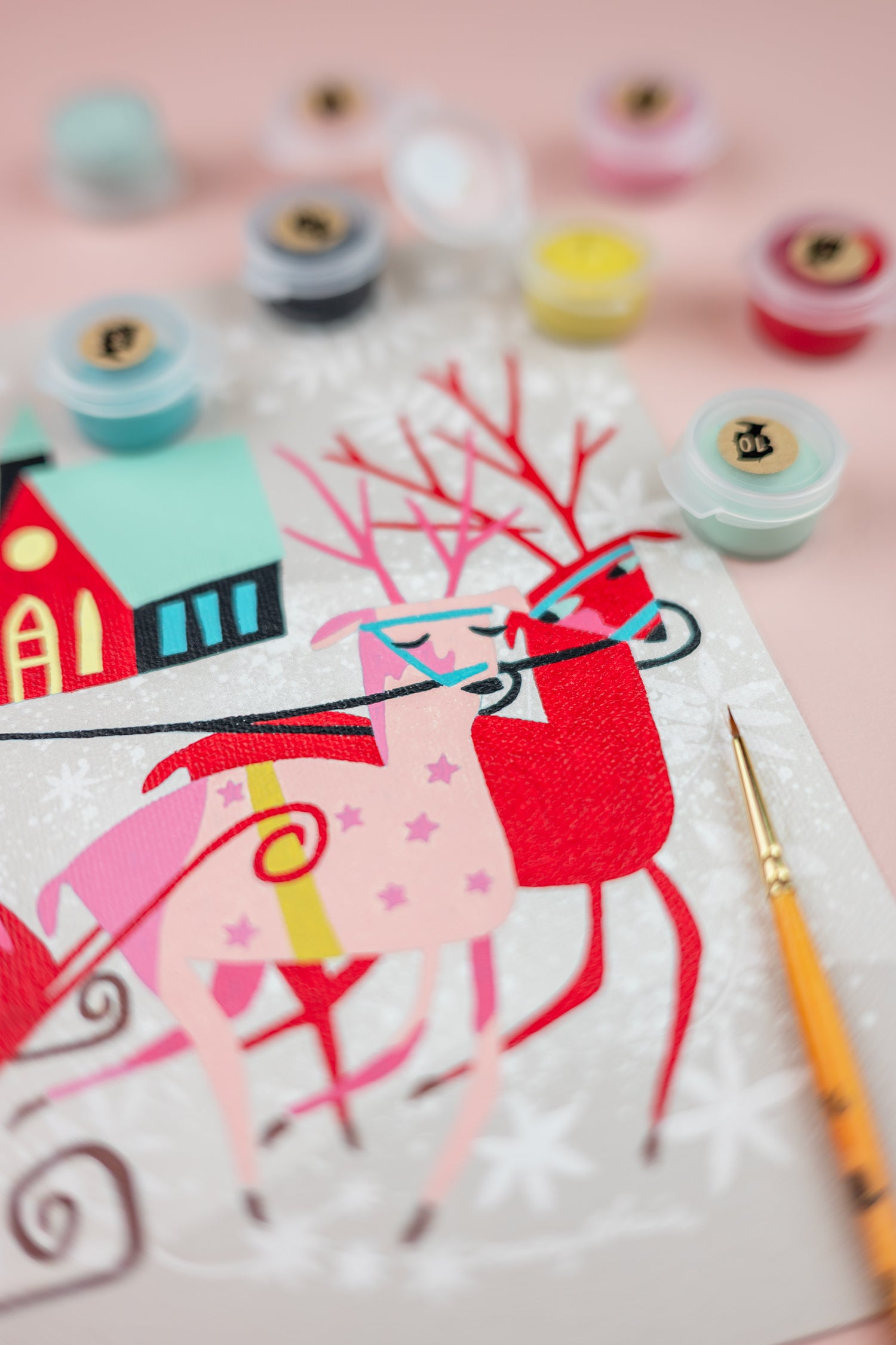 Colorful paint-by-number kit of a reindeer pulling a sleigh with paint containers and a brush on a pink background.