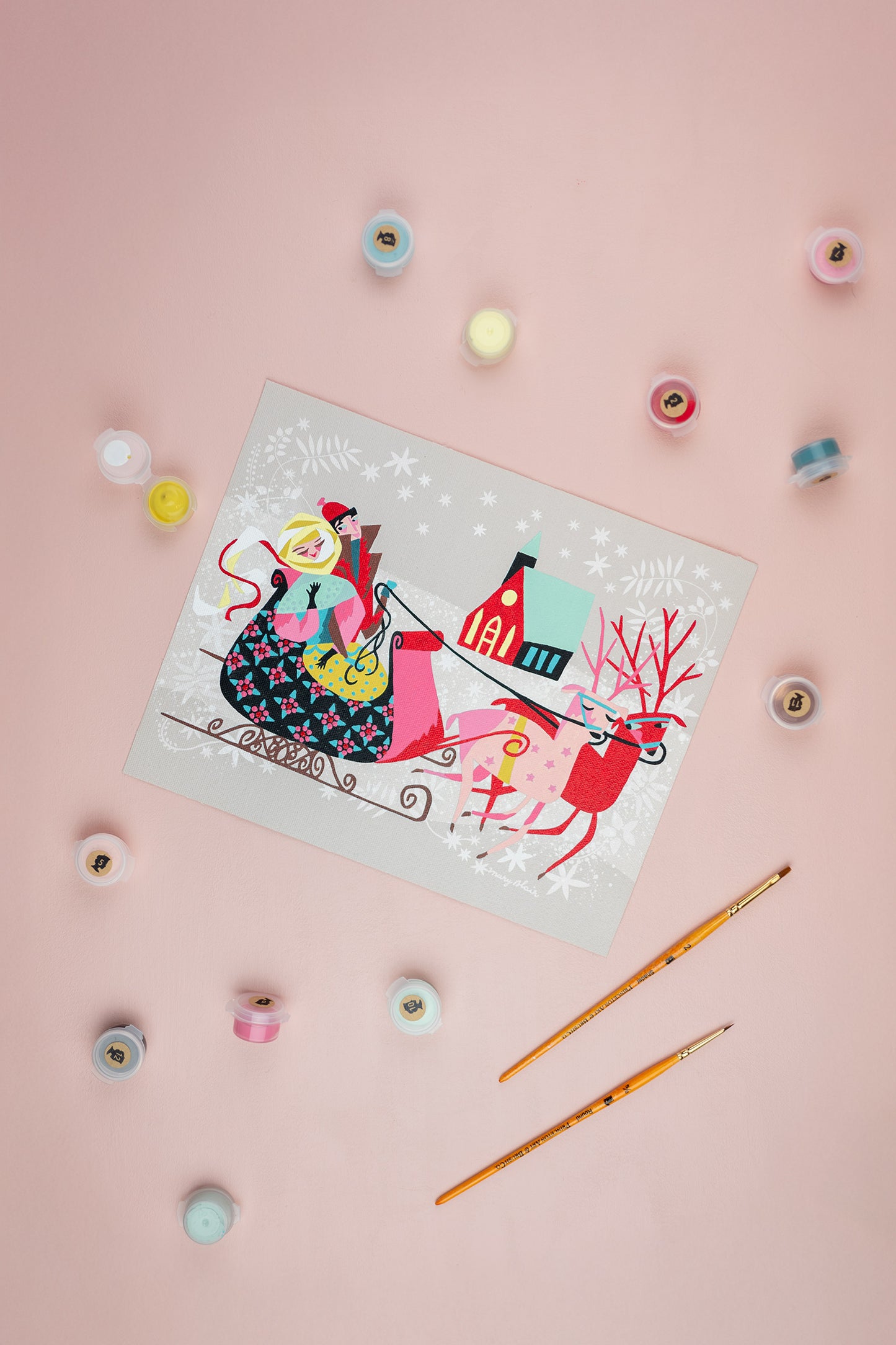 Colorful paint-by-number kit of a reindeer pulling a sleigh with a couple inside. Scatttered around it are paint containers and a brush on a pink background.