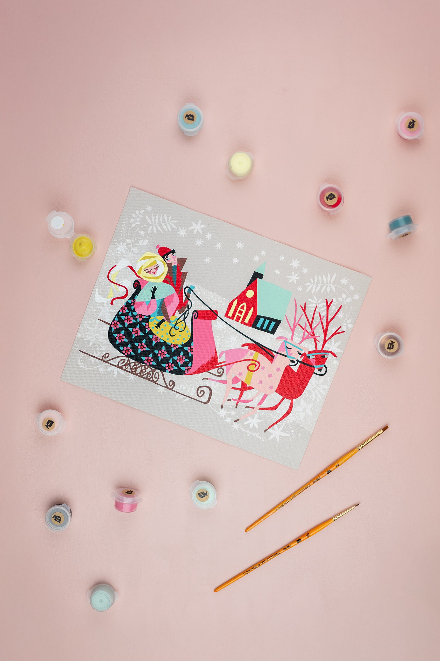 Colorful paint-by-number kit of a reindeer pulling a sleigh with a couple inside. Scatttered around it are paint containers and a brush on a pink background.
