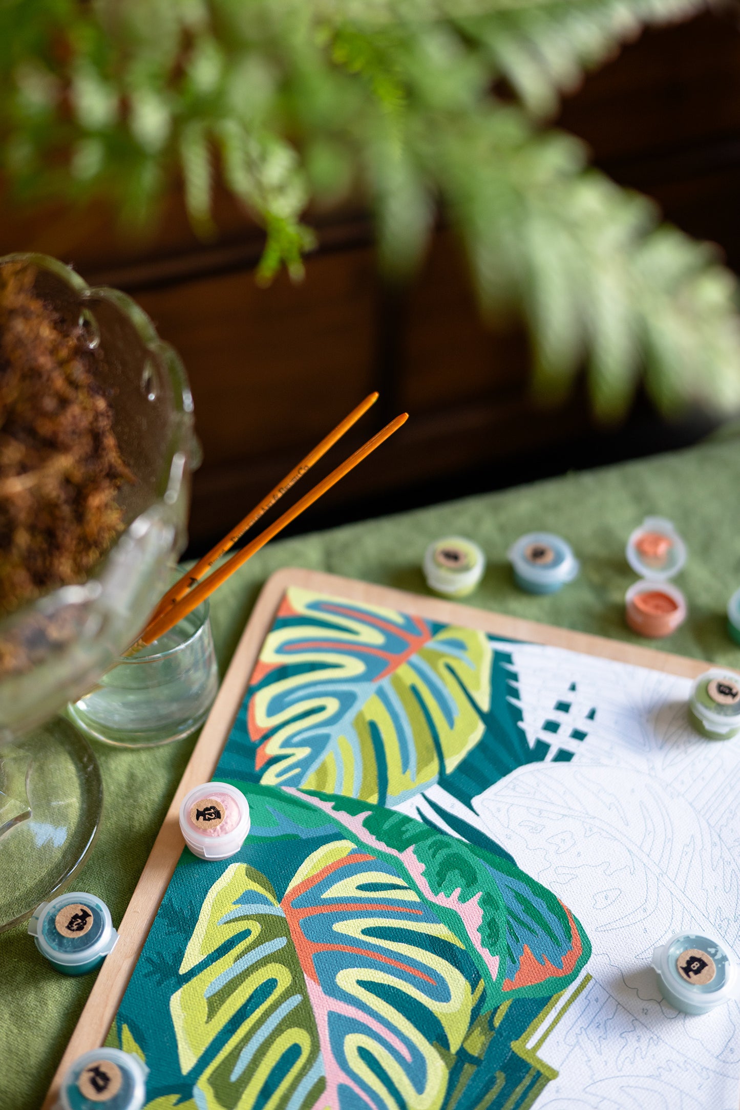 Vintage Verde paint-by-number kit in progress with tropical leaf artwork, paint pots, and brushes on a green table beside houseplants.
