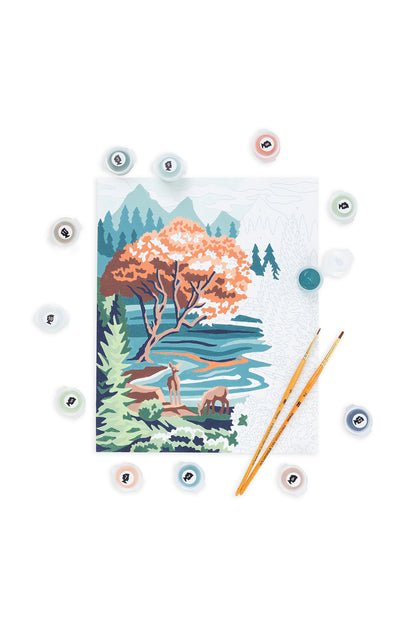 Partially painted Pine & Lake paint-by-number canvas on a white background, surrounded by numbered paint pots and two paintbrushes. The artwork shows a Pacific Northwest lake scene with pine trees, an autumn-colored tree, flowing water, and deer, with some sections still unpainted.
