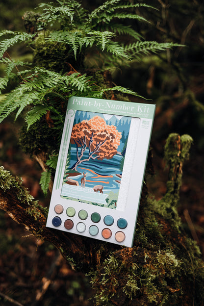 Pine & Lake paint-by-number kit by Elle Crée resting on a mossy tree branch, featuring a Pacific Northwest lake and pine forest illustration with paint pots included.