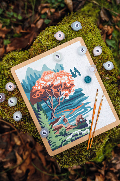 Half-painted Pine & Lake paint-by-number kit with visible unpainted sections, shown outdoors on moss with paint pots, brushes, and a Pacific Northwest lake scene.