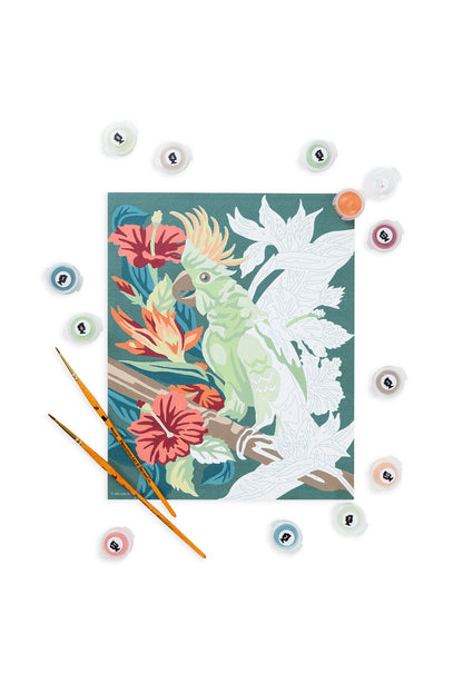 Partially painted Perched in Paradise cockatoo paint-by-number kit showing tropical flowers on an 8x10 canvas, surrounded by acrylic paint pots and two detail brushes.