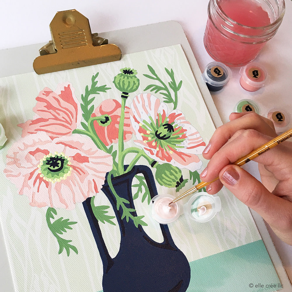 A hand is holding a paintbrush that is dipped in a pink paint pot on top of a poppies paint-by-number kit from Elle Crée.